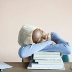 Sleeping African American female student in headscarf sitting at table with opened notebook and lying on stack of textbooks