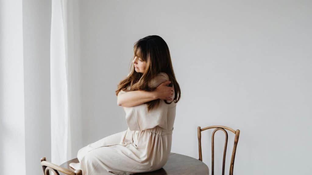 Woman in a light dress sits on wooden table, embracing as a sign of self-care