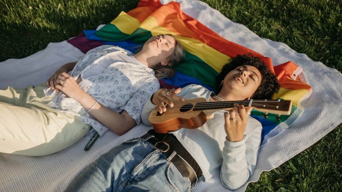 Two individuals embracing love and relaxation, lying on a rainbow flag with a ukulele, outdoors.