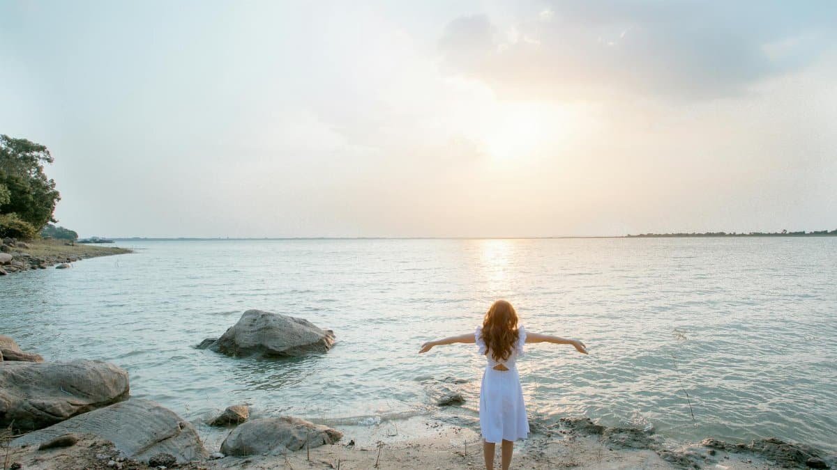 A woman in a white dress stands on a rocky beach, embracing the serene seascape at sunset.