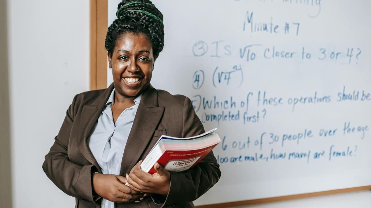 A smiling teacher holding textbooks stands confidently in front of a whiteboard.