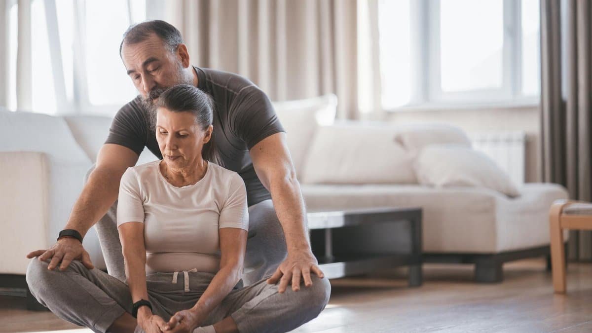 Elderly couple engaging in a yoga session indoors, practicing bound angle pose with focus and support.