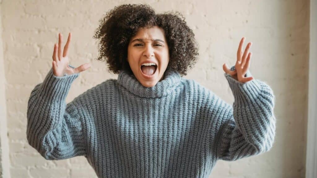 Irritated African American female with raised arms looking at camera while shouting loudly near wall in light room at home