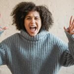 Irritated African American female with raised arms looking at camera while shouting loudly near wall in light room at home