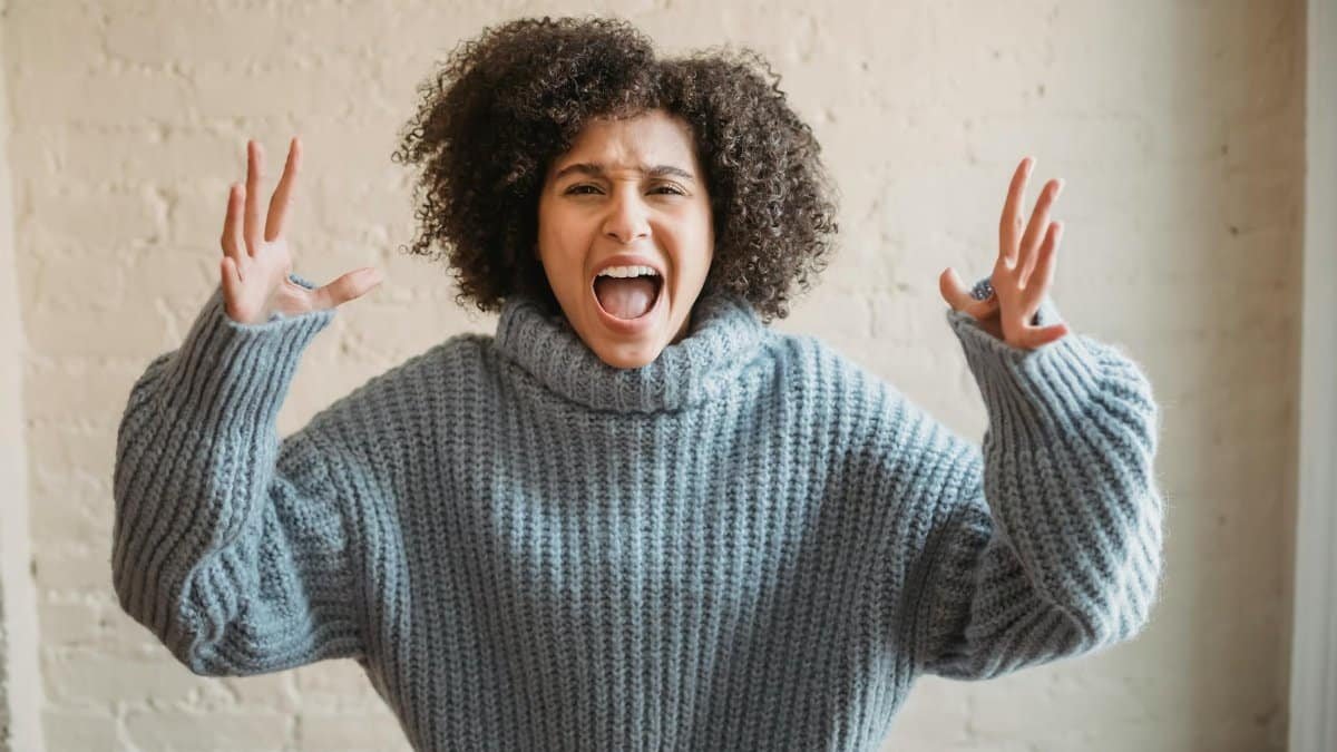 Irritated African American female with raised arms looking at camera while shouting loudly near wall in light room at home