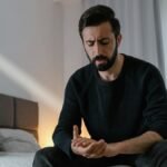 A worried man in a black sweater sits on a bed in a bright room, expressing stress.