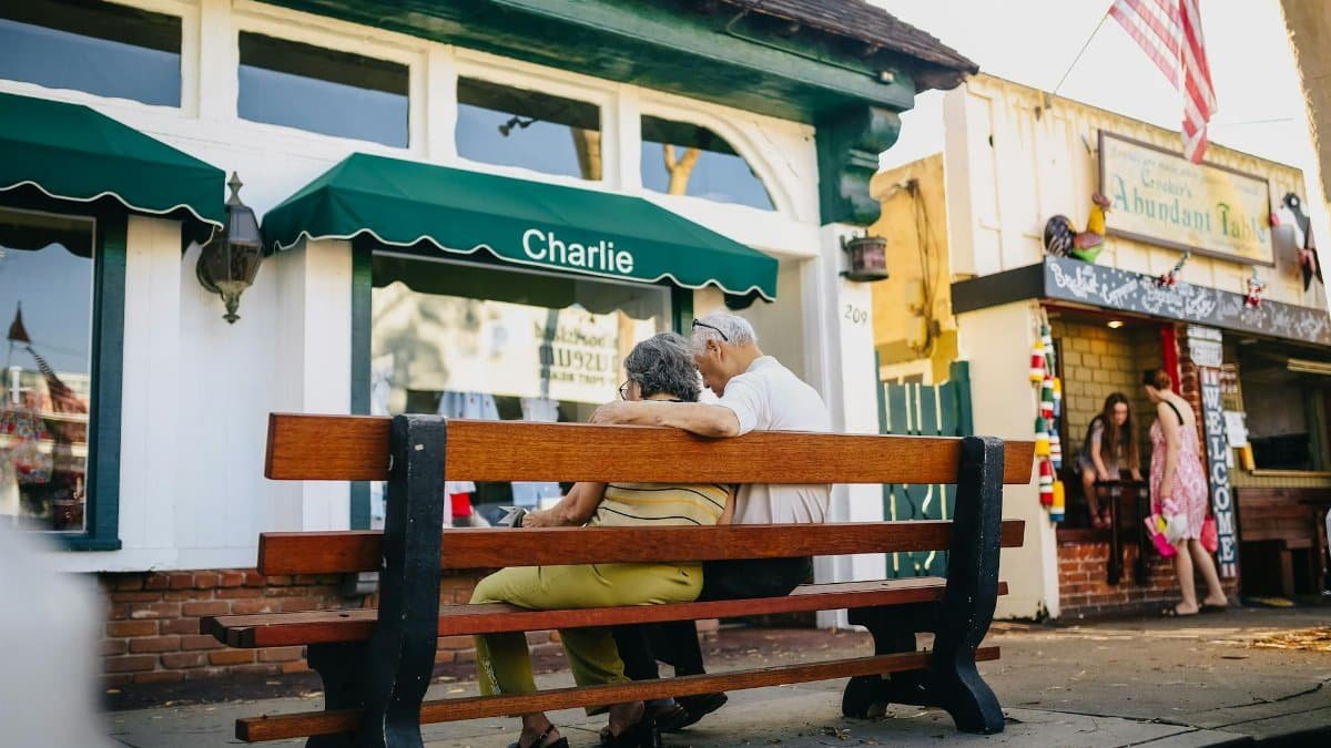A senior couple sits together on a bench outside a shop, embracing under an American flag.