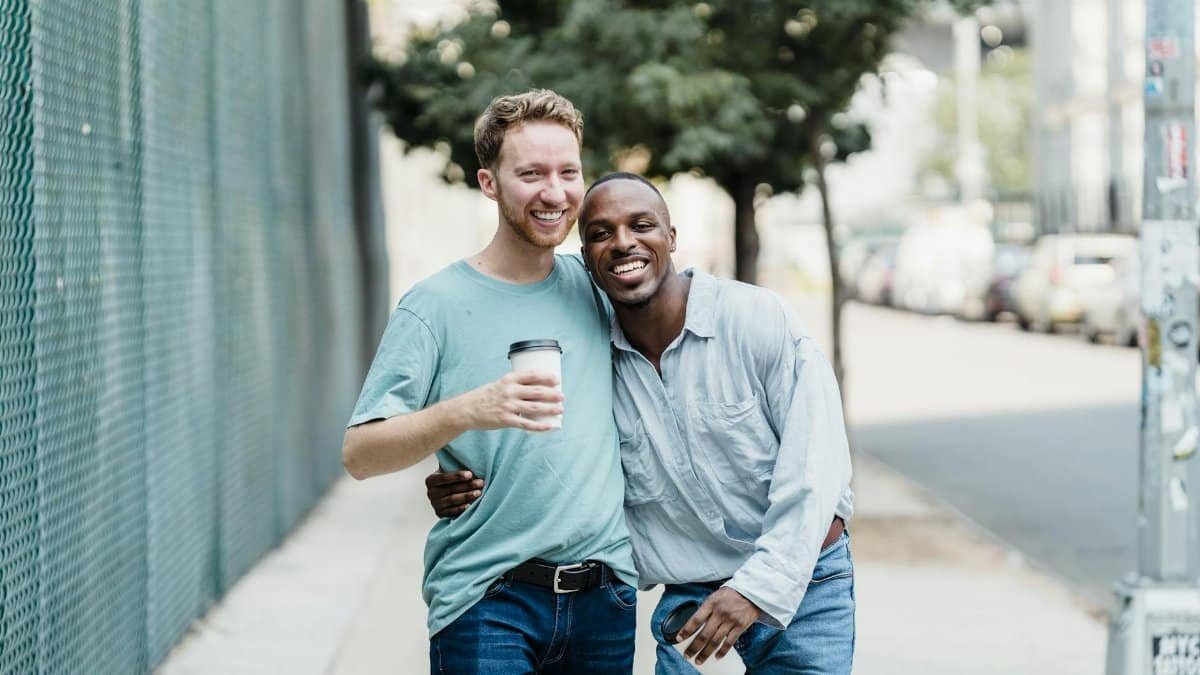 Two men walking with coffee, smiling and embracing on a sunny city sidewalk.