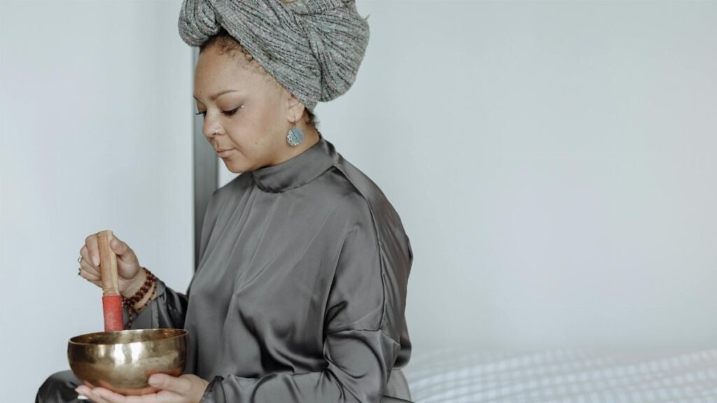 Woman in gray attire meditating indoors with a singing bowl for relaxation.