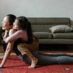 A woman practicing yoga with her daughter in a cozy living room, embracing and smiling.