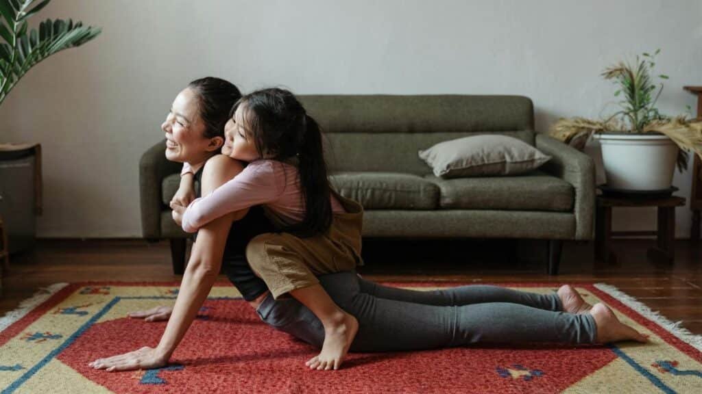 A woman practicing yoga with her daughter in a cozy living room, embracing and smiling.