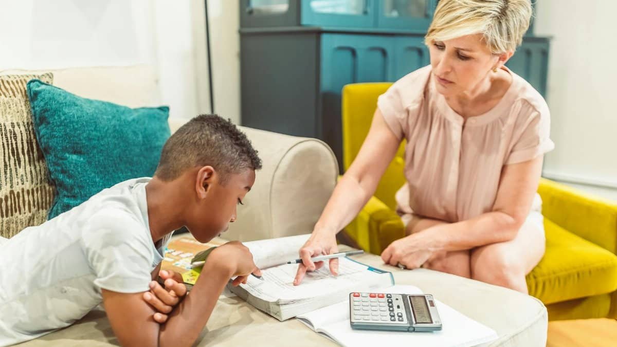 Caucasian woman and African American boy studying together on a sofa with books and a calculator.