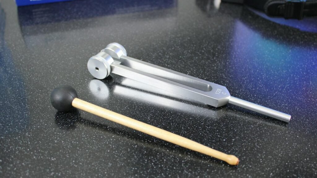 A shiny metal tuning fork with mallet on speckled black table, for sound therapy or music.