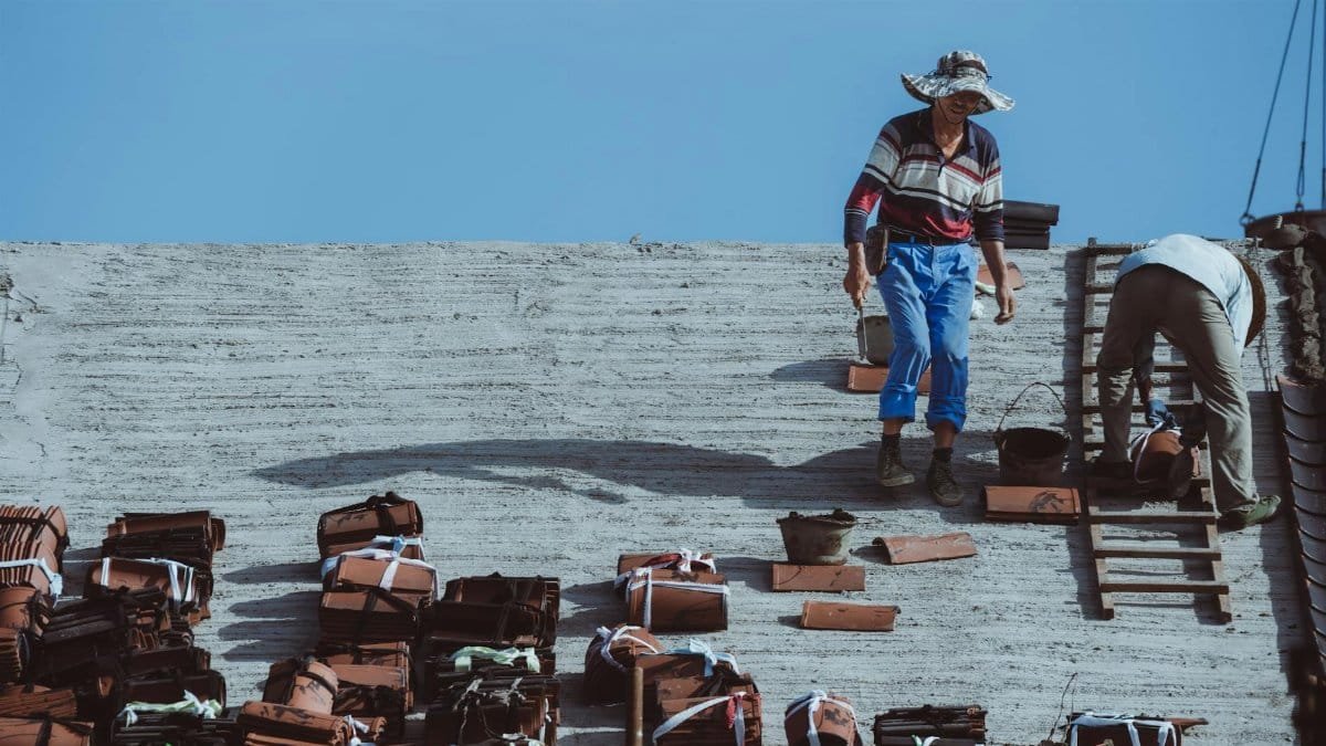 Two construction workers installing roof tiles on a building under a clear blue sky.