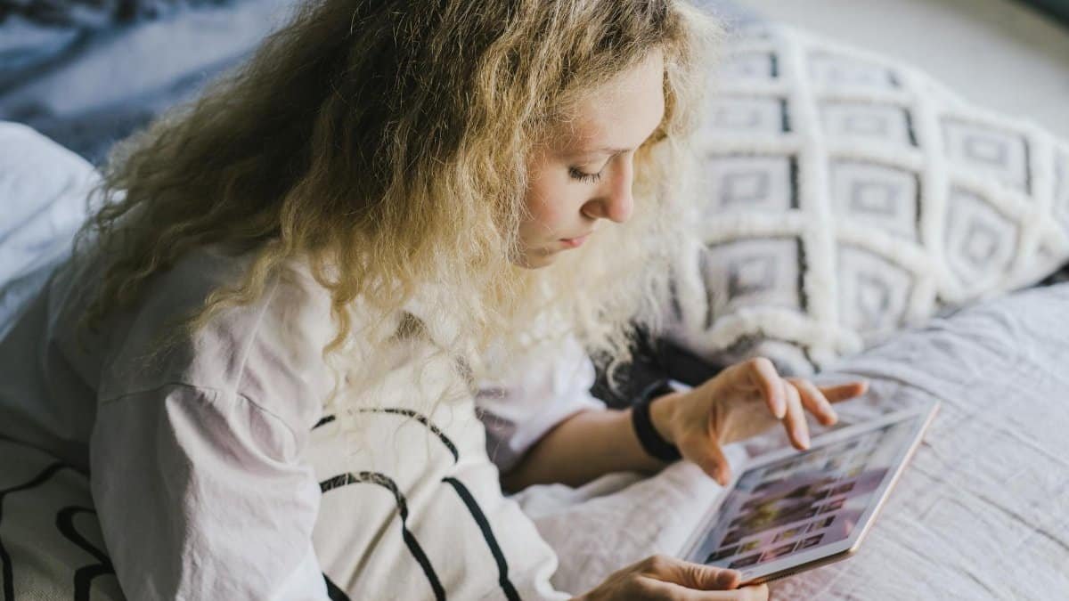 A young woman sits on her bed scrolling on a tablet, showcasing modern relaxation and technology.