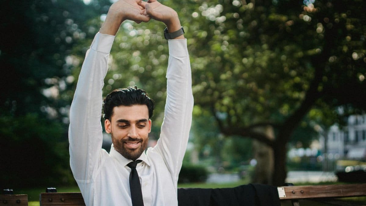 Smiling businessman stretches arms while sitting on park bench, wearing white shirt and tie.