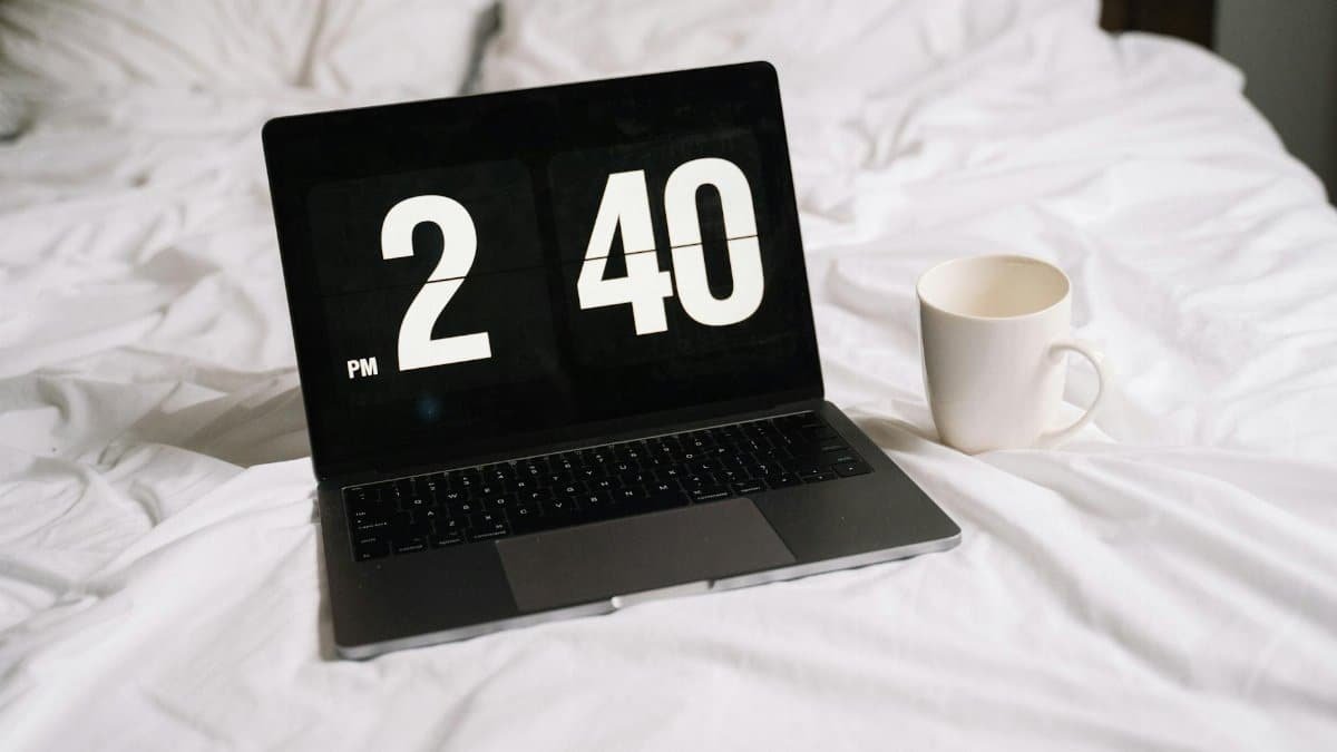A laptop displaying time and a coffee mug on a white bed, creating a relaxed work or leisure atmosphere.