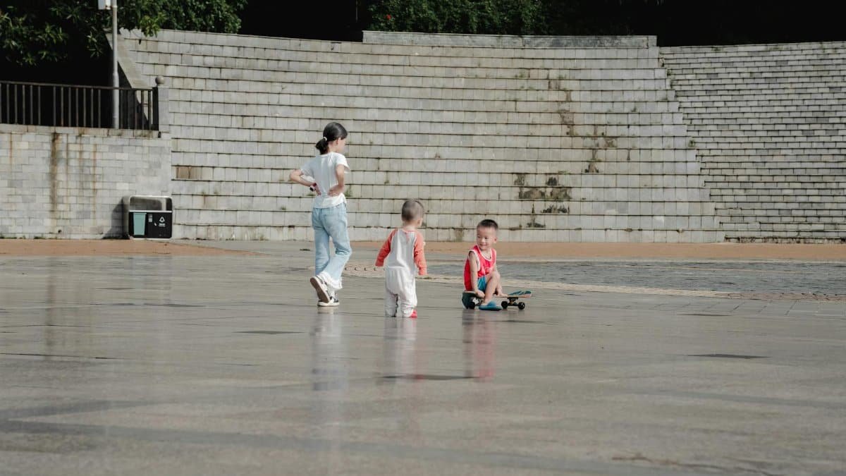Three children enjoying outdoor playtime on a skateboard at a public square during daytime.