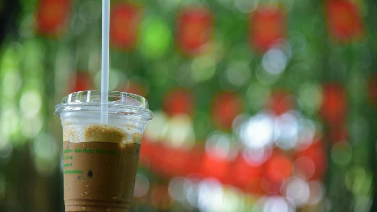 A close-up of iced coffee in a clear cup with green and red bokeh background outdoors.