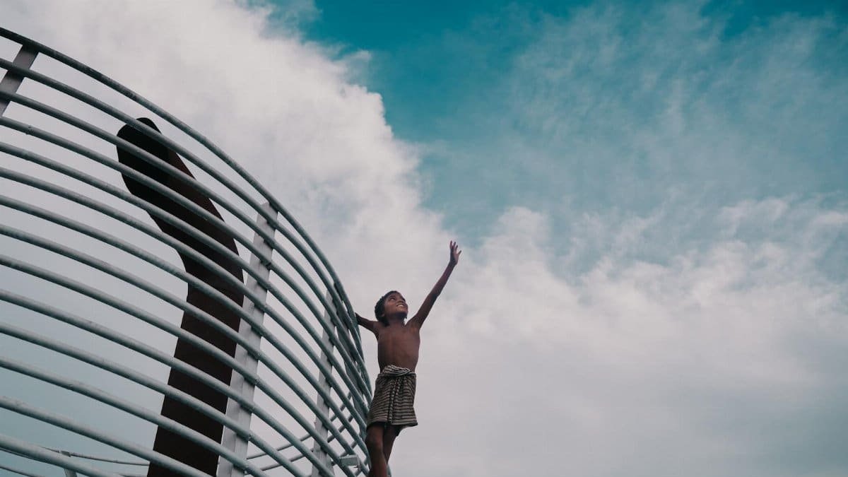 Young boy standing on metal structure, reaching up towards the sky with clouds in the background.