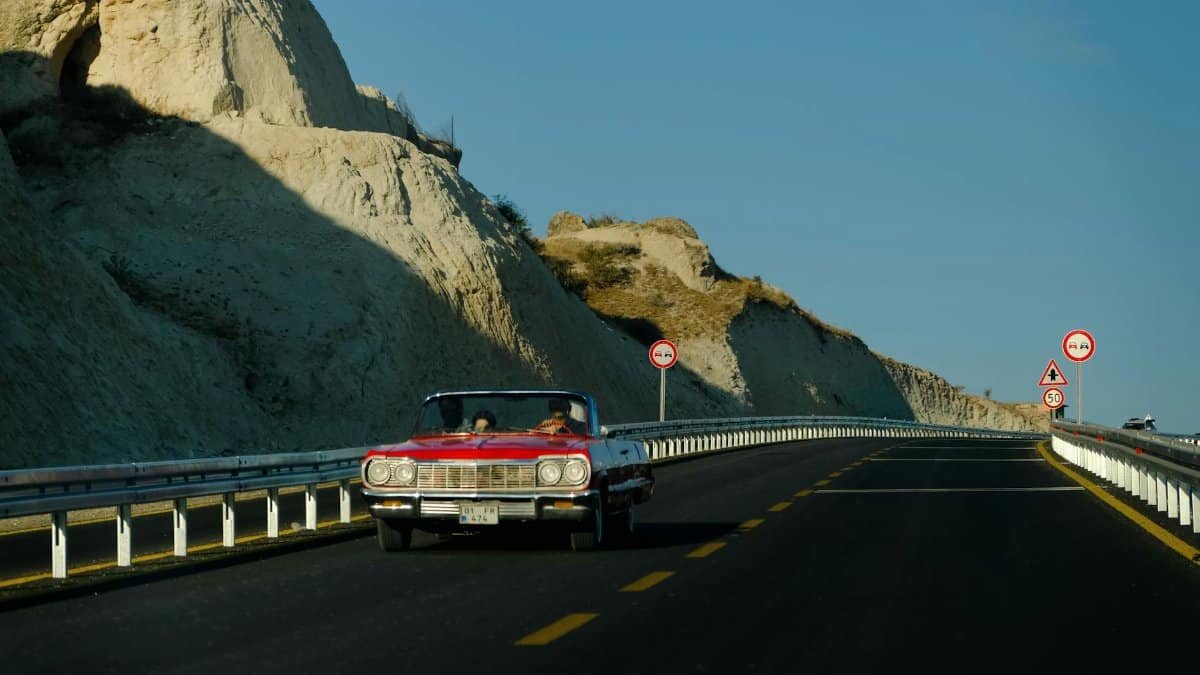 A red vintage convertible car driving on a scenic highway with rocky cliffs under a clear blue sky.