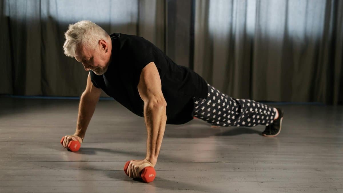 Senior man performing a push-up using dumbbells for strength training indoors.