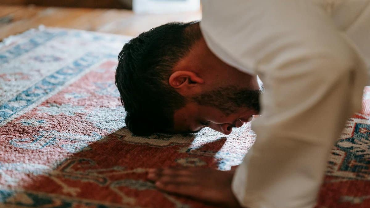 A Muslim man performing a prayer ritual inside on a traditional rug.