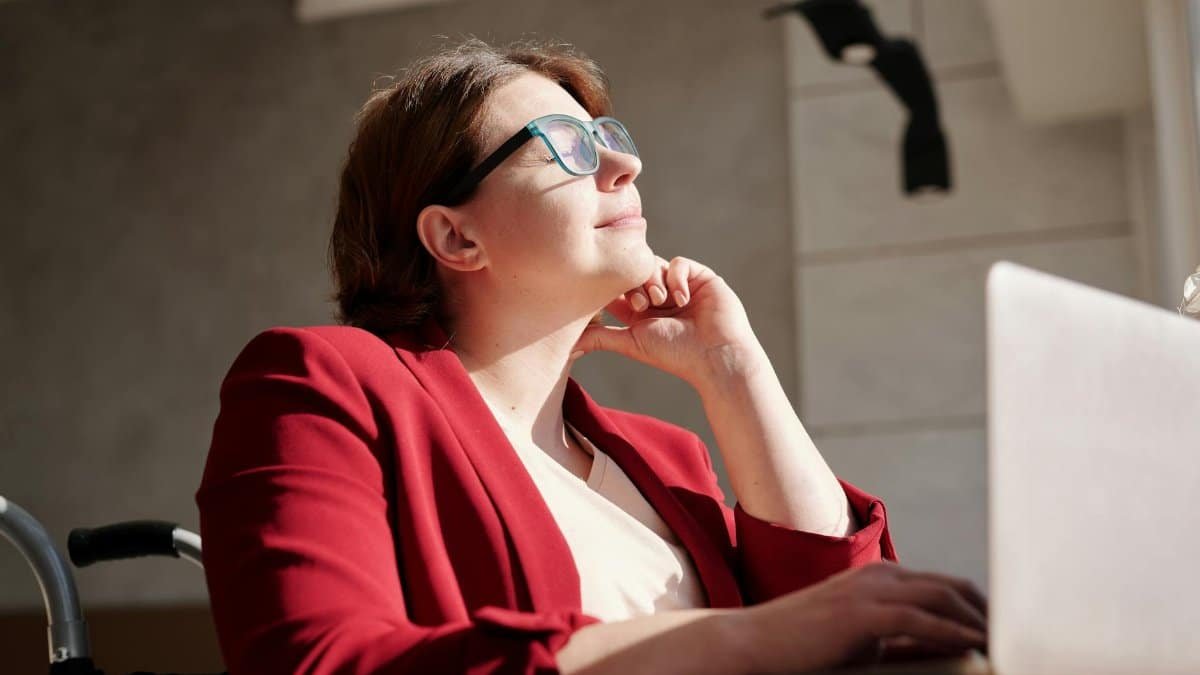 Woman in wheelchair wearing red suit and glasses, working on a laptop in a bright café.