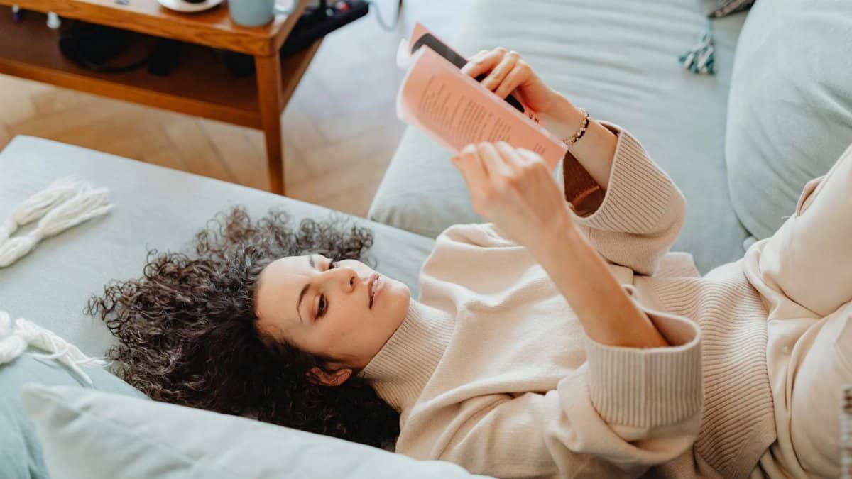 Woman in sweater reading a book on a cozy sofa, enjoying leisure time indoors with a cup of tea.