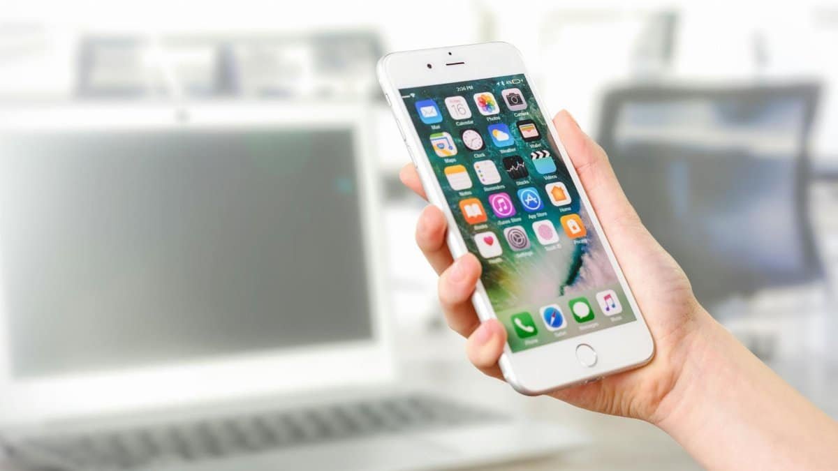 Close-up view of a smartphone displaying apps, held by a hand, with a blurred laptop in the background.