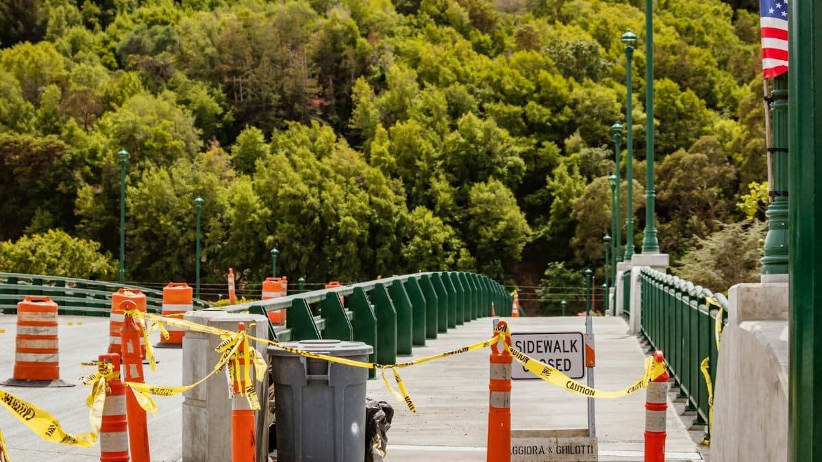 Orange construction cones and caution tape block a bridge sidewalk.
