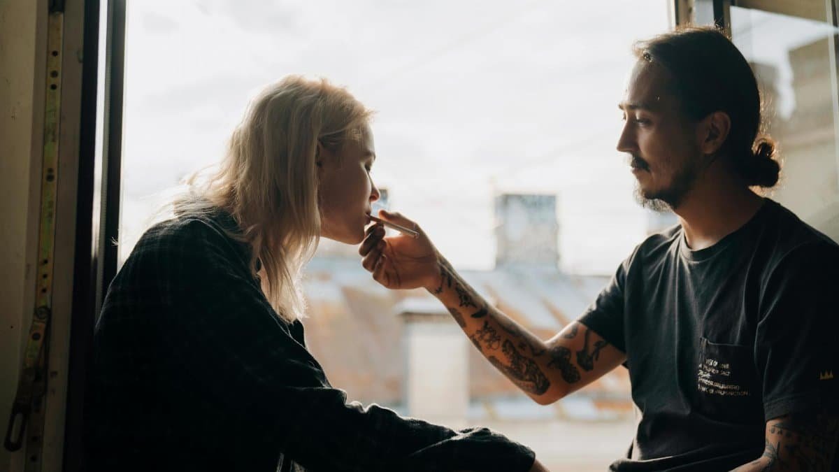 A young couple shares a moment by the window as they smoke together, enjoying a bonding time.