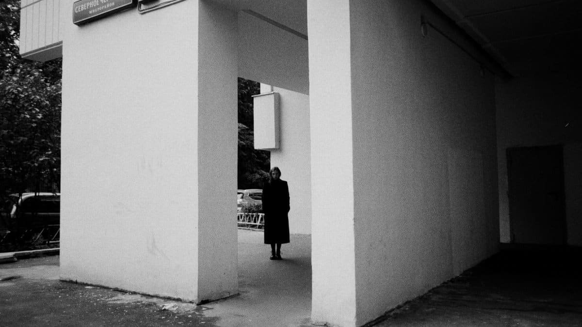 A solitary figure stands in a stark urban passageway, bathed in contrast.