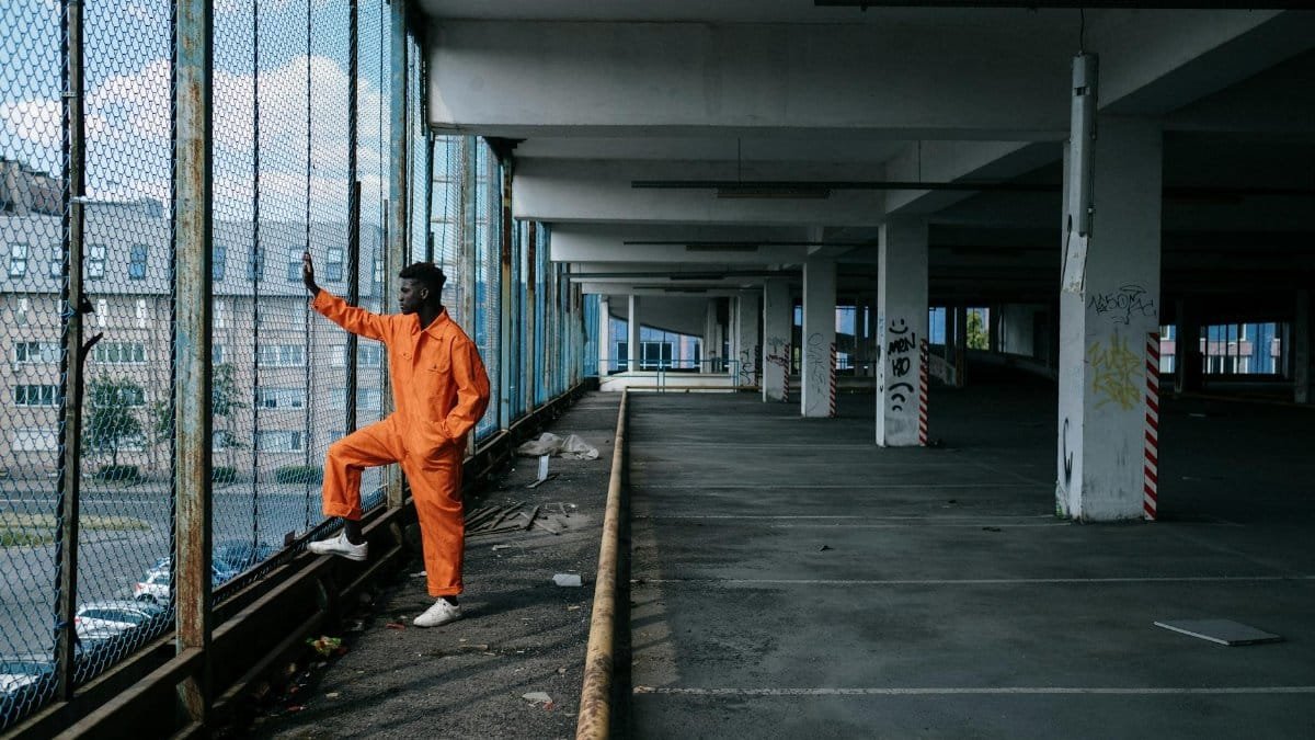A man in orange overalls stands by a wire fence in a parking garage. Urban setting.