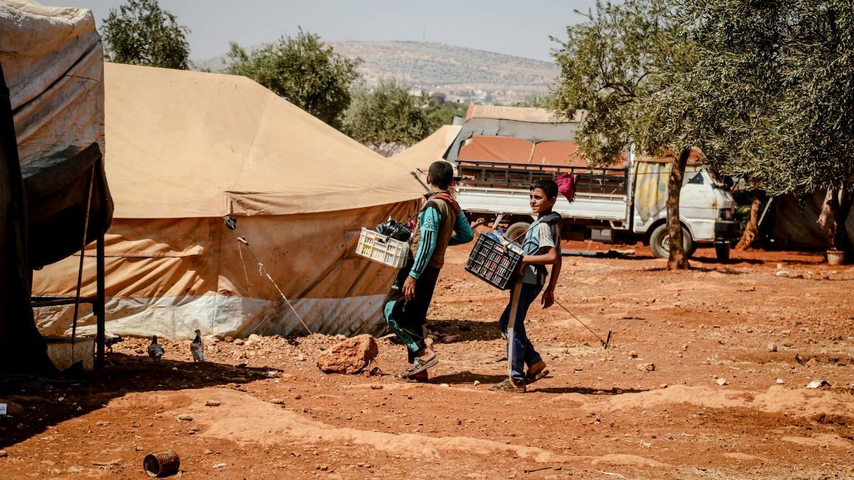 Two boys carrying crates walk through a refugee camp in Idlib, Syria, amidst tents and trees.