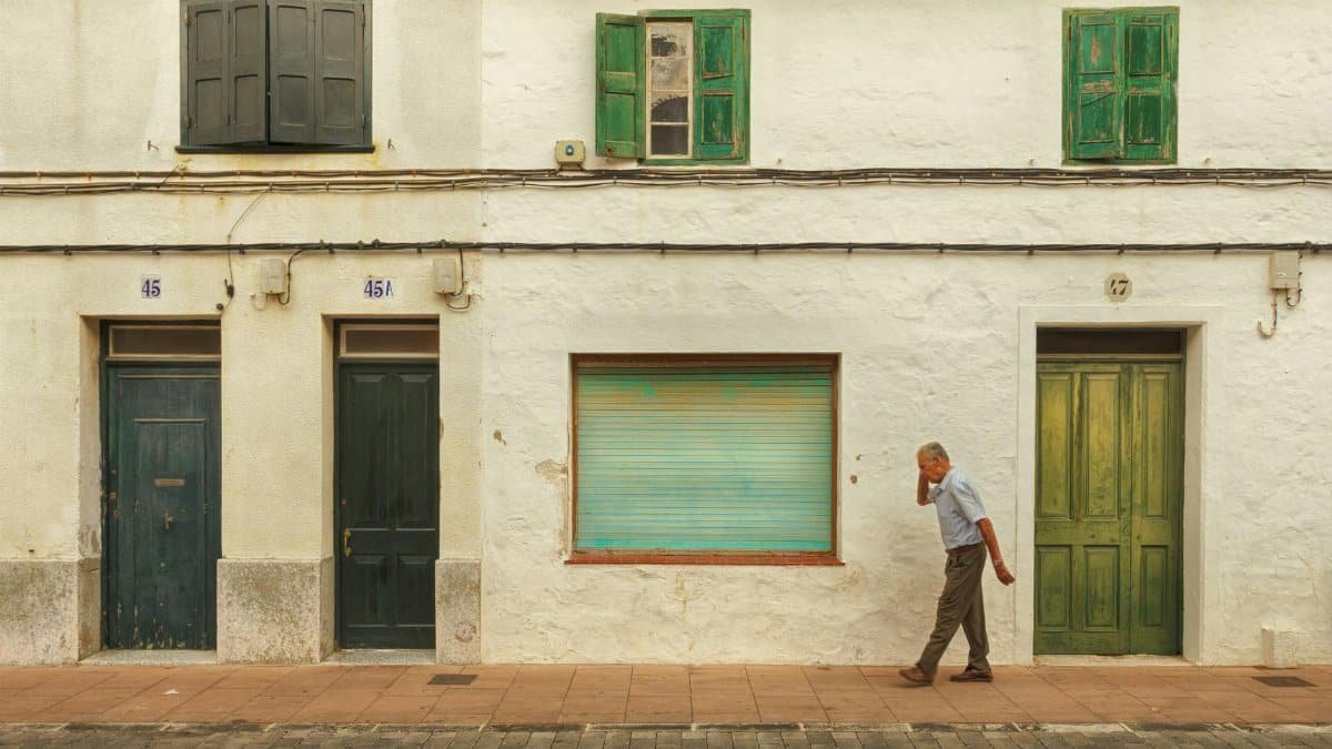 An elderly man walks past a white urban building with green doors and shutters.