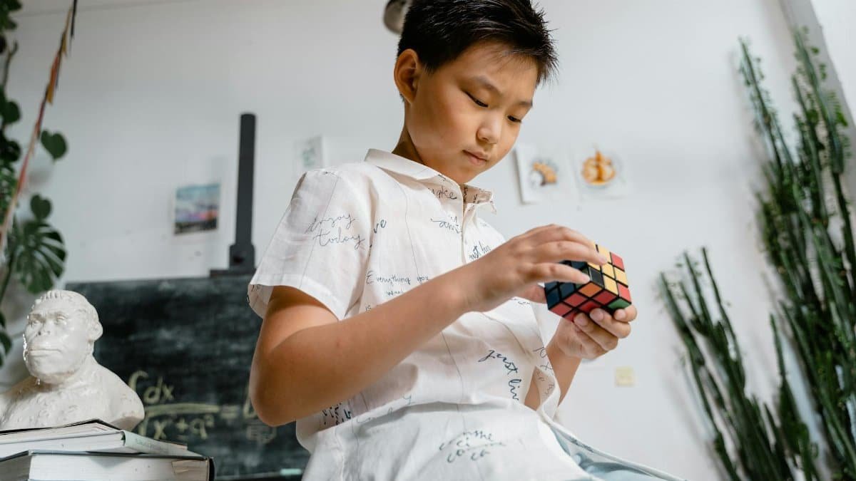 Asian boy thoughtfully solving a Rubik's Cube in a classroom, surrounded by books and a classroom setting.
