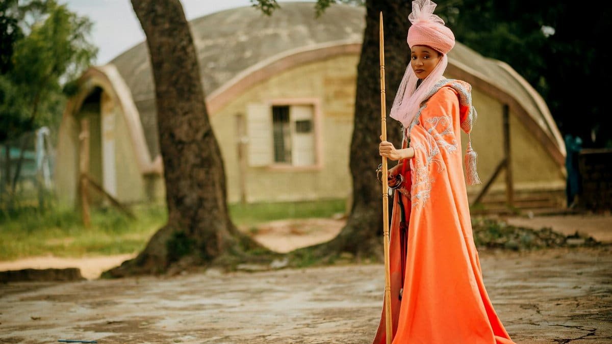 Woman in vibrant traditional attire posing outdoors with a stick in hand.