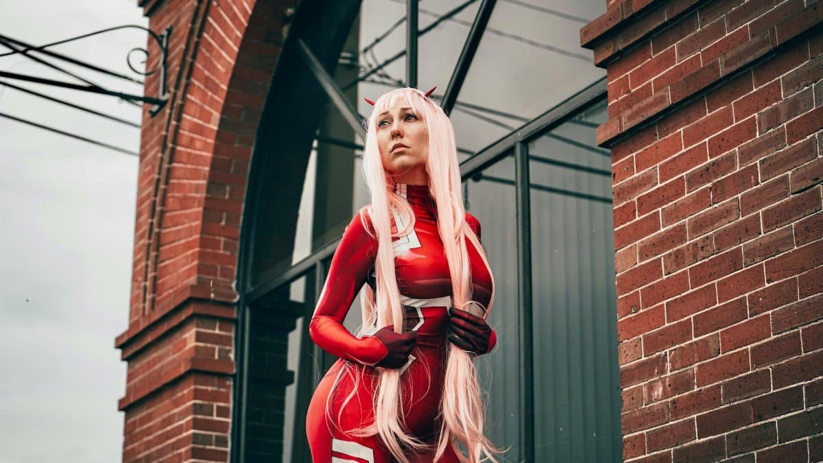 A cosplayer in a red costume poses confidently against a brick building.