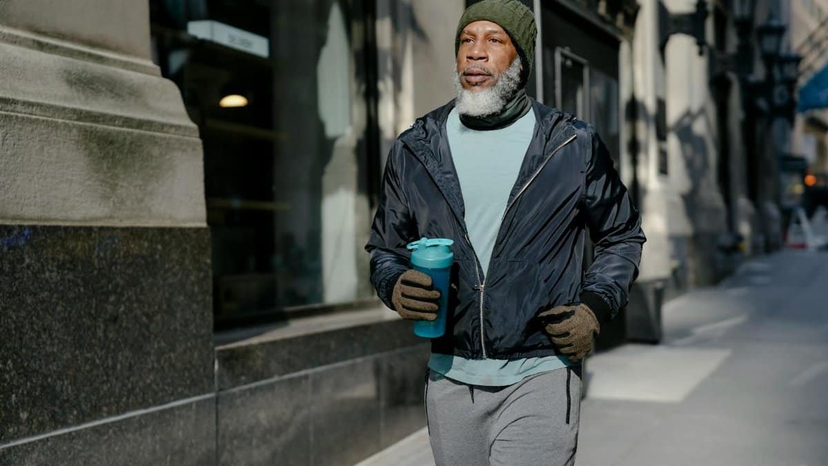 Serious elderly African American male in activewear and knitted hat and gloves running on pavement near building with sport bottle