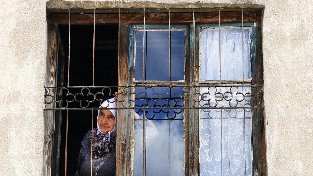 An elderly woman looking out through an old barred window of a weathered building.