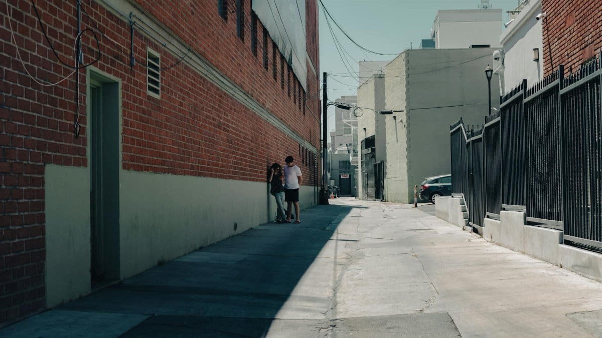 A couple stands in a shadowy urban alley beside a brick building on a sunny day.