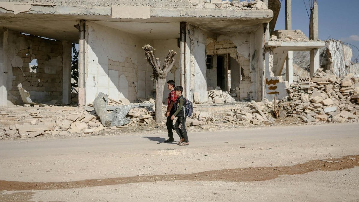 Two boys walk along a road beside a damaged building in Idlib, Syria, showcasing resilience.