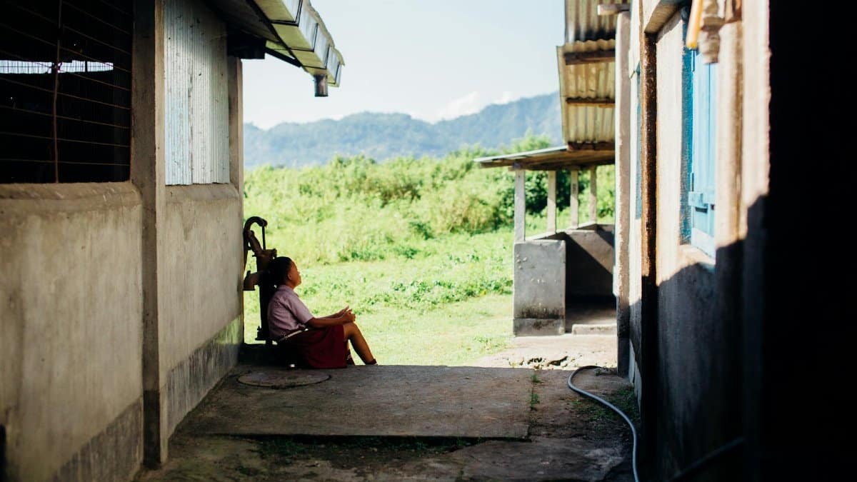 A woman in a skirt relaxes by a rustic building with mountains in view, under daylight.