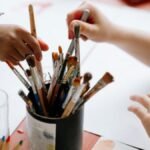 Hands reaching for paintbrushes in an artistic indoor setting.
