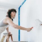 A woman stands on a ladder painting a wall white, part of a DIY home renovation.