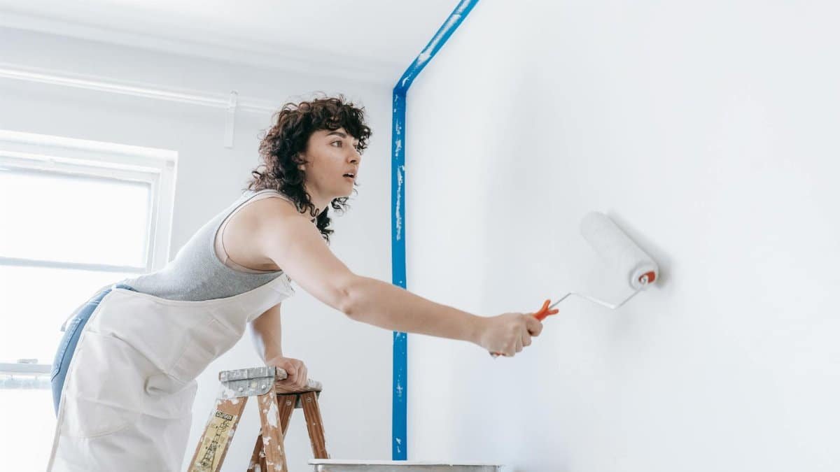 A woman stands on a ladder painting a wall white, part of a DIY home renovation.