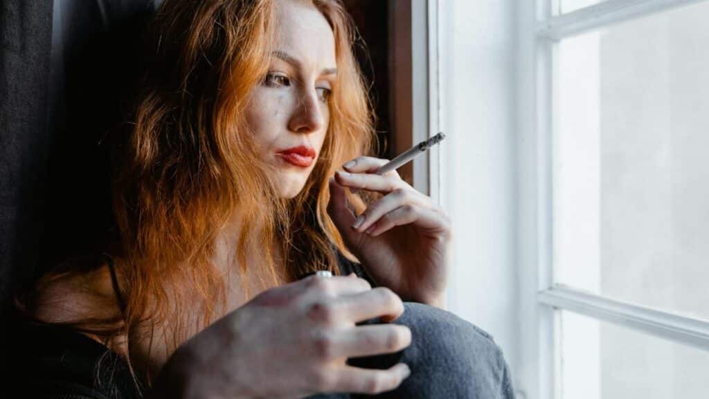 Pensive young woman with red hair holding a cigarette by a window, reflecting indoors.