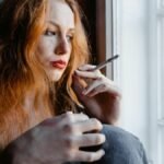 Pensive young woman with red hair holding a cigarette by a window, reflecting indoors.