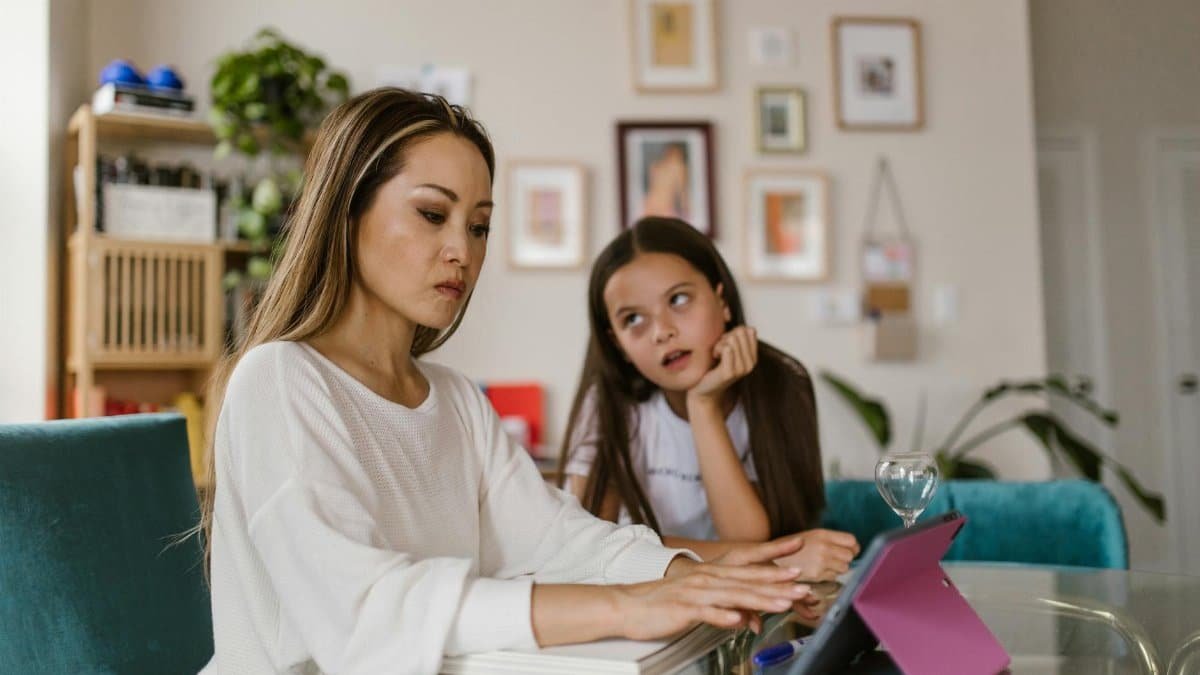 A mother working on her tablet at home while her daughter watches from nearby, illustrating remote work challenges.